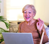 Join One of Our Virtual Groups Happy mature woman waving to someone while having a video call over laptop at home. Gray-haired senior woman waving hand in front of laptop while having video call with her family members.